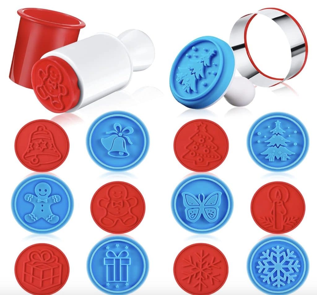 A set of colorful cookie stamps perfect for creating the best Christmas cookies. Red and blue stamps feature festive images like a bell, tree, gingerbread man, butterfly, candle, gift, and snowflakes. A metal circular cutter and red pusher complete this cheerful collection.